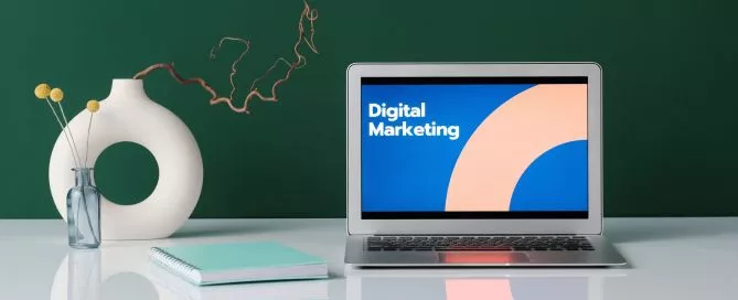 Computador em cima de uma mesa com "Marketing Digital" escrito na tela.