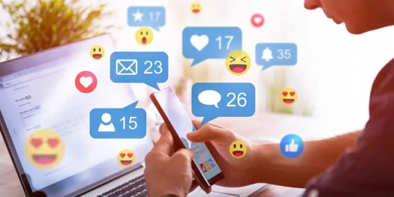 Uma pessoa mexe em um computador e há animações coloridas de ícones de redes sociais como emojis e notificações com likes, comentários e compartilhamentos